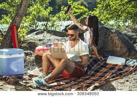 Young Couple Having Picnic At Riverside In Sunny Day. Woman And Man Spending Time On The Nature Toge