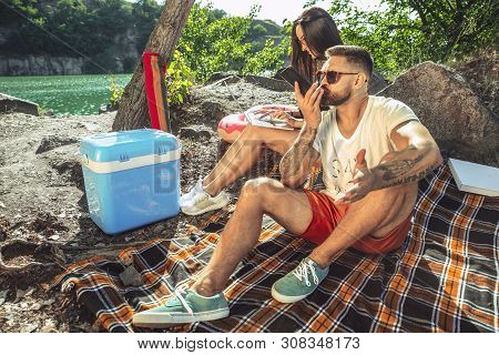 Young Couple Having Picnic At Riverside In Sunny Day. Woman And Man Spending Time On The Nature Toge