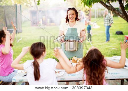 Playful Little Girls Are Sitting At The Picnic Table And Are Cheering For Their Mother Who Is Bringi