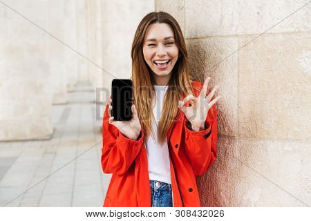 Cheerful attractive young girl wearing raincoat spending lovely time outdoors, showing blank screen mobile phone, ok