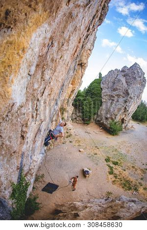 Athletic Man Climbs A Rock, 7c Category Route In The Forest With Rope, Against Sunset And The Other 