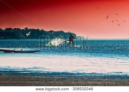 Silhouette Of A Surfer Folds Sail On The Beach Against The Backdrop Of The Seascape And Dramatic Sun