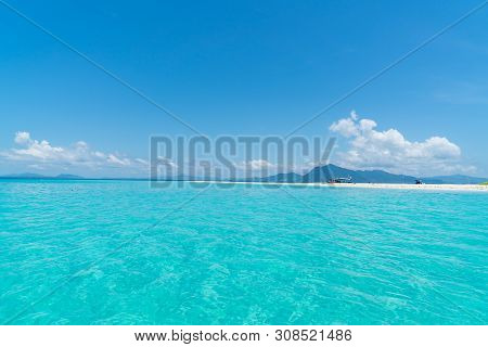 Idyllic Tropical Sea, Sand Bar And Distant Mountains Under Blue Sky With Few Puffy White Clouds.