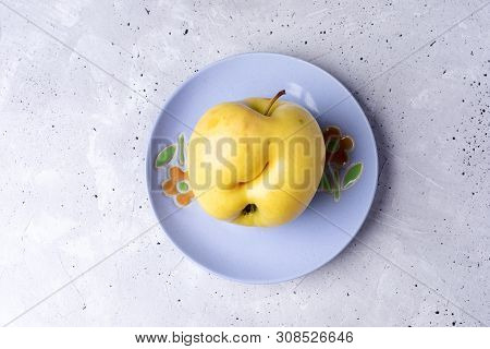 One Ugly Yellow Apple On Lilac Plate On Grey Concrete Background.
