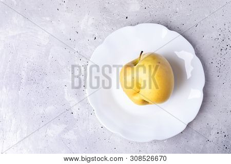 One Ugly Yellow Apple On White Figured Plate On Grey Concrete Background.