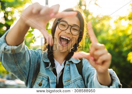Image of a cheerful screaming smiling cute young student girl wearing eyeglasses outdoors in nature park make photo frame gesure.