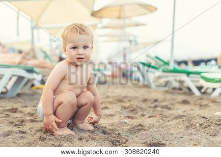 Cute Caucasian Toodler Boy Squatting Alone On Sandy Beach Between Chaise-lounge. Adorable Happy Chil