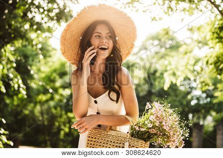 Image of excited happy young pretty woman outoors in green park walking in hat talking by mobile phone.