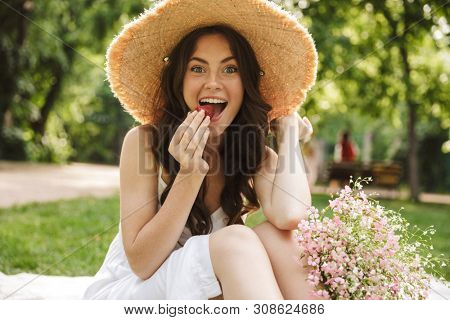 Image of excited happy young pretty woman outoors in green park in hat sitting on grass eat strawberry.