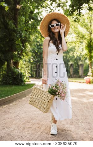 Image of smiling happy young pretty woman outoors in green park walking in hat.