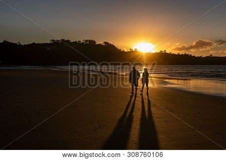 A Couple Walking An Empty Oneroa Beach, Waiheke Island, New Zealand In A Golden Sunset