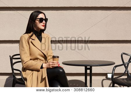 Outdoor Shot Of Stylish Woman Wearing Fashionable Beige Coat, Having Break In Street Outdoor Cafe An
