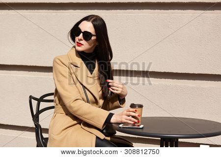 Horizontal Shot Of Young Beautiful Brunette Woman In Stylish Sunglasses And Beige Coat Sitting At Ta