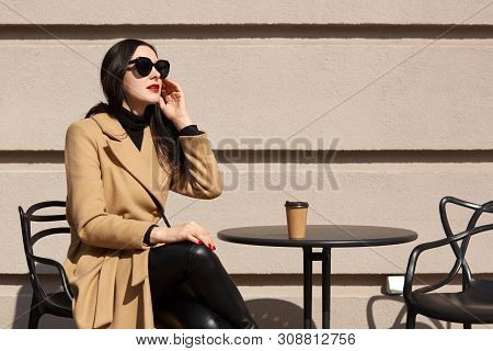 Outdoor Shot Of Brunette Woman Wearing Fashionable Coat, Black Outfit And Sunglasses, Sitting At Res