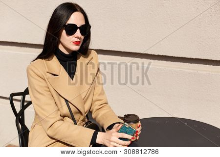 Image Of Young Beautiful Brunette Woman In Black Sunglasses And Beige Stylish Coat Sitting At Table 