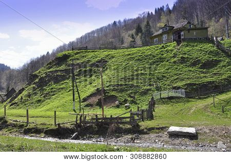 A House In The Mountains With Outbuildings On A Green Hill Is Fenced With A Wooden Fence. In The Bac