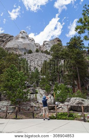 A Young Man Stands By A Railing At The Base Of The Mountain To Take A Picture Of Mount Rushmore With