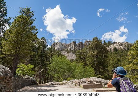 A Young Man Stands At The Base Of The Mountain To Take A Picture Of Mount Rushmore With Pine Trees A