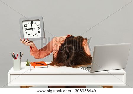 Young Exhausted Tired Woman With Lowered Head Holding Square Clock, Sit And Work At Desk With Pc Lap