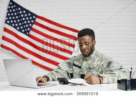 Male Soldier Or Worker Of American Army Sitting At Table And Writing Documents, Using Laptop. Americ
