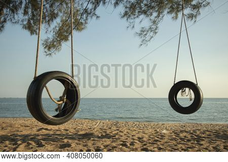Tire Swings Hang From Trees On The Sandy Beach. With A Background Of Blue Sea And Sky With Coluds