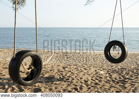 Tire Swings Hang From Trees On The Sandy Beach. With A Background Of Blue Sea And Sky With Coluds