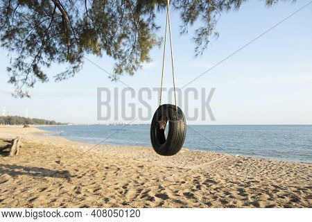Tire Swings Hang From Trees On The Sandy Beach. With A Background Of Blue Sea And Sky With Coluds