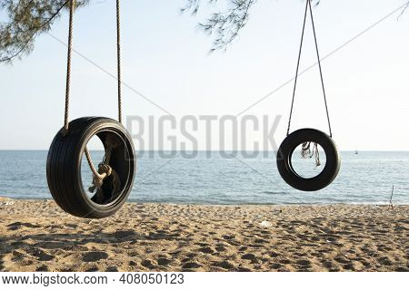 Tire Swings Hang From Trees On The Sandy Beach. With A Background Of Blue Sea And Sky With Coluds
