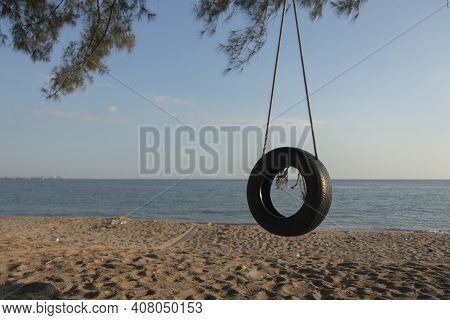 Tire Swings Hang From Trees On The Sandy Beach. With A Background Of Blue Sea And Sky With Coluds