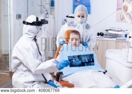 Little Girl And Mother Wearing Ppe Suit Looking At Dental Radiography In Dentist Office During Covid