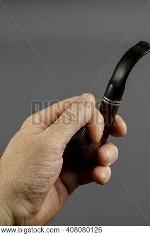 Hand Holds A Smoking Pipe On A Gray Background. Bend A Briar Or Heather Root Smoking Pipe. Tobacco S