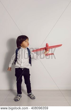 Boy Child In A White Shirt And Jeans Flies On A Red Airplane Toy At The White Wall Of The House