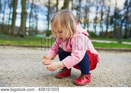 Adorable Baby Girl In Park, Sitting On Hunkers And Playing With Little Stones. Little Child Having F