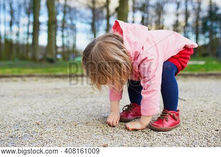 Adorable Baby Girl In Park, Sitting On Hunkers And Playing With Little Stones. Little Child Having F