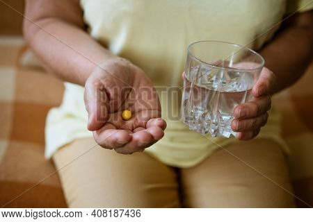 Mature Woman With A Glass Of Water Holds A Tablet In Her Hand While Sitting On The Couch. Treatment 
