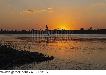 Silhouette Of Tropical River Bank At Dusk Sunset Sun In Orange Sky With Traditional Sailing Felluca 