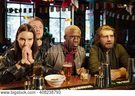 Group of young intercultural friends in casualwear standing by bar counter with drinks and snacks while watching football broadcast in pub