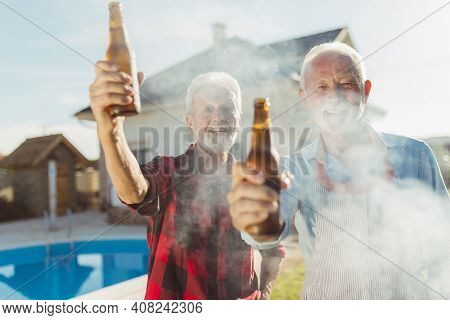 Two Senior Men Making A Toast With Bottles Of Beer While Grilling Meat At Backyard Barbecue Party By