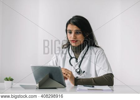 Indian Female Doctor In A White Coat With A Stethoscope Conducts An Online Consultation In Her Offic