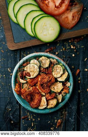 high angle view of some slices of dried tomato and zucchini, served as appetizer or snack in a rustic green plate, and some slices of raw tomato and zucchini, on a dark gray rustic wooden surface