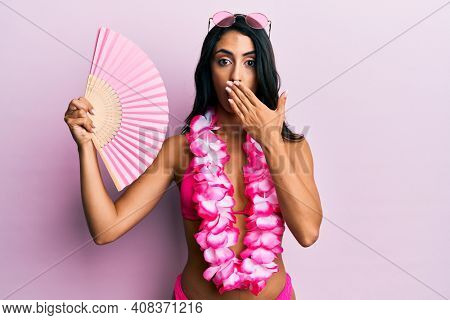 Beautiful hispanic woman wearing bikini holding handfan covering mouth with hand, shocked and afraid for mistake. surprised expression 
