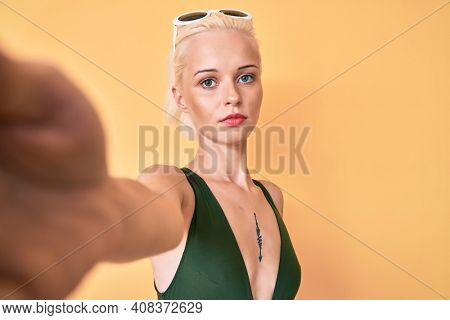 Young blonde woman with tattoo wearing swimwear taking a selfie thinking attitude and sober expression looking self confident 