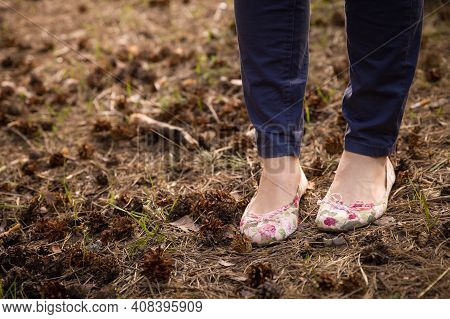 Closeup Of Legs Of A Young Woman In Jeans And Pumps. Dry Spruce Needles And Cones Lie On The Ground.