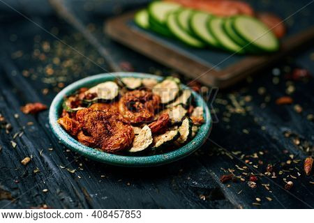 a rustic green plate with some slices of dried tomato and zucchini, served as appetizer or snack, on a dark gray rustic wooden surface next to some slices of raw tomato and zucchini