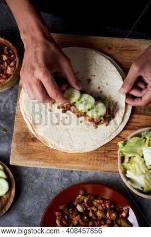 high angle view of a young caucasian man filling a durum or a burrito, with chicken meat cooked with different vegetables such as onion or red and green pepper, and fresh lettuce and raw cucumber