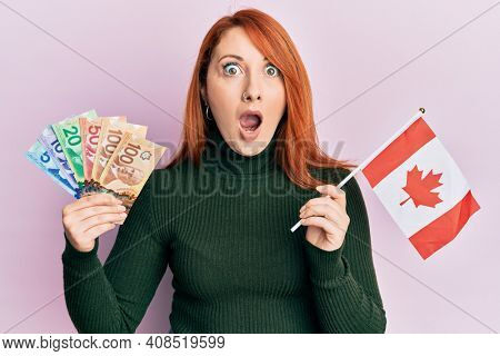 Beautiful redhead woman holding canadian dollars and canada flag afraid and shocked with surprise and amazed expression, fear and excited face. 