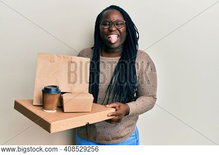 Young black woman with braids holding take away food sticking tongue out happy with funny expression. 