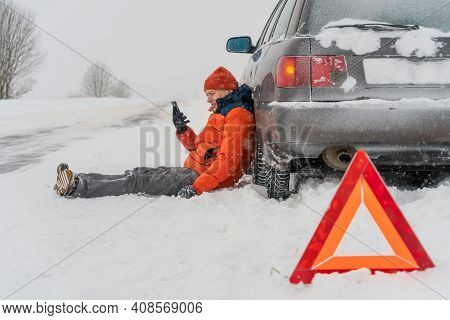 Desperate Young Man In An Orange Jacket Sits Near A Car And Talks On The Phone On A Snowy Road.he Is