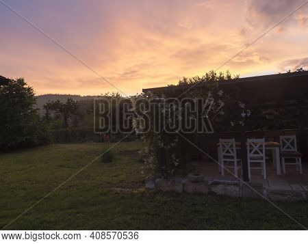 View Of Summer Garden With Tiber Wooden Gazebo Pergola With Blooming Climbing Plant Flowers And Whit