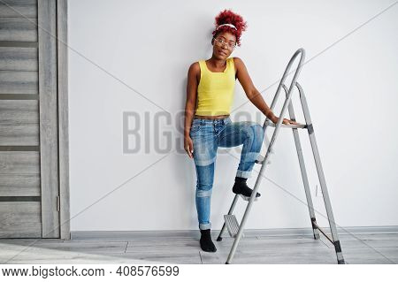 African American Woman In Yellow Singlet And Eyglasses Against White Wall Stand Om Iron Ladder.